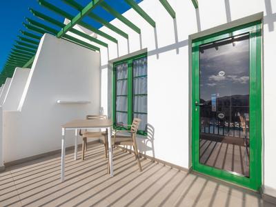 Balkon mit weißer Wand, grünem Türrahmen, Tisch und zwei Stühlen bei Sonnenschein.