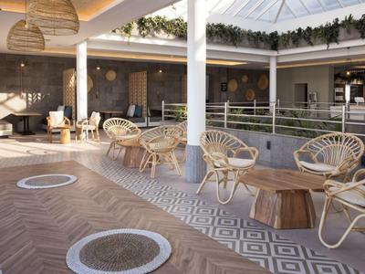 Heller Loungebereich mit Korbsesseln, Holztischen, Pflanzen und geometrischem Bodenmuster.