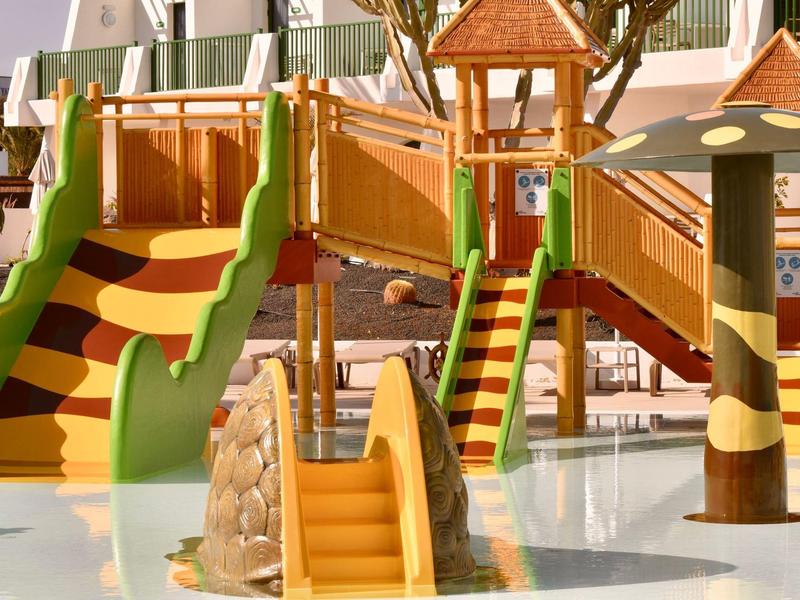 Bunter Kinderspielplatz mit Rutschen, Holzbrücken und pilzförmigen Spielgeräten auf weichem Boden.