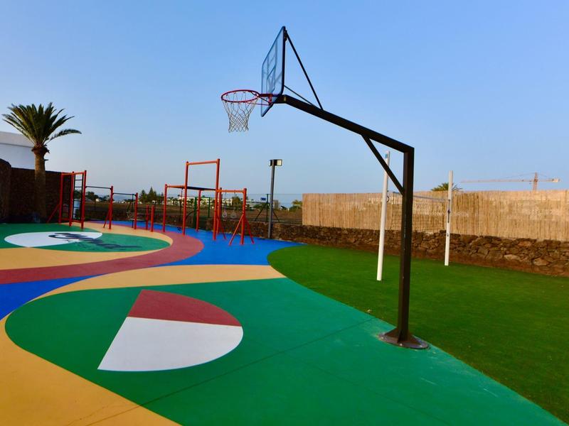 Bunter Basketballplatz mit grünem, blauem und gelbem Boden, Korb und Spielgeräten im Hintergrund.