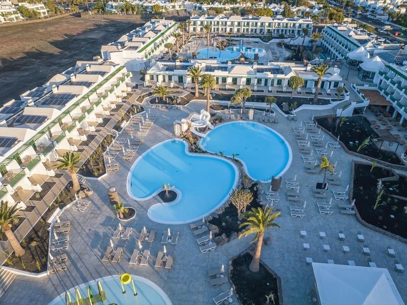 Großer Hotelkomplex mit zwei verbundenen, blauen Schwimmbädern und zahlreichen Sonnenschirmen.