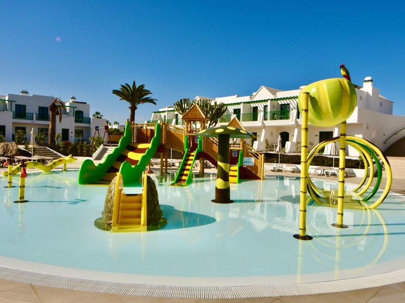 Kinderpool mit bunten Wasserrutschen, Wasserspielen und Palmendekoration vor Hotelgebäuden.