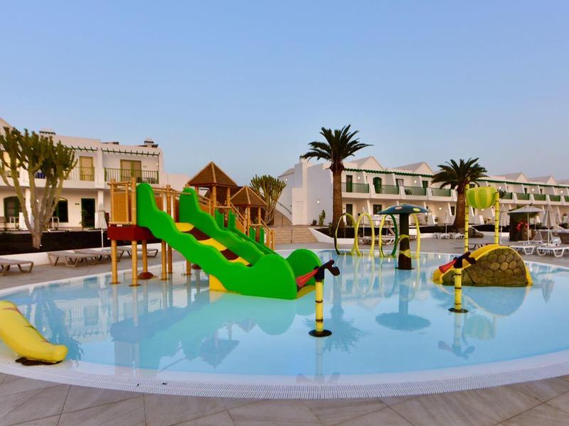 Rundlicher Pool mit bunten Rutschen und Wasserspielen vor weißen Gebäuden bei klarem Himmel.