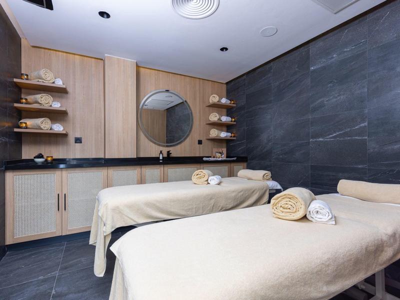 Moderner Massageraum mit zwei Liegen, hellen Holzmöbeln und dunklen Steinwänden.