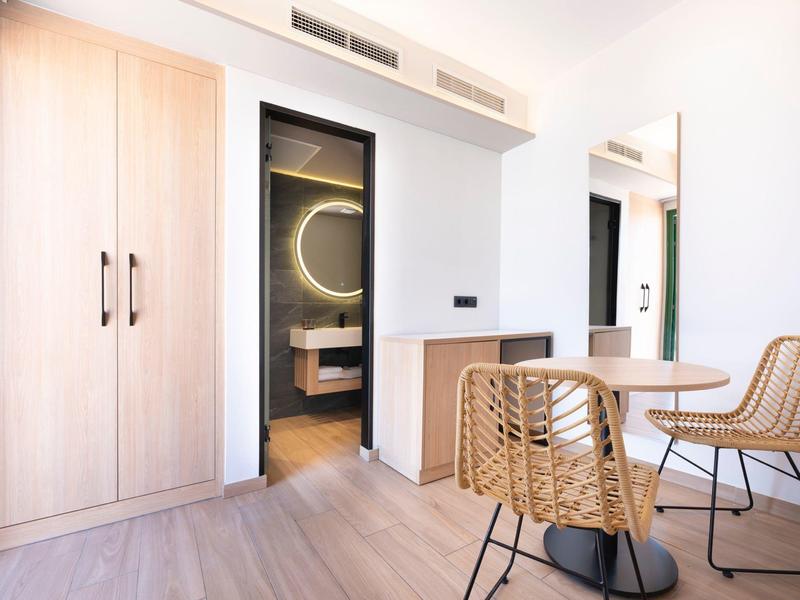 Modern eingerichteter Hotelraum mit Holzschrank, Stühlen und einem Badezimmer mit beleuchtetem Spiegel.