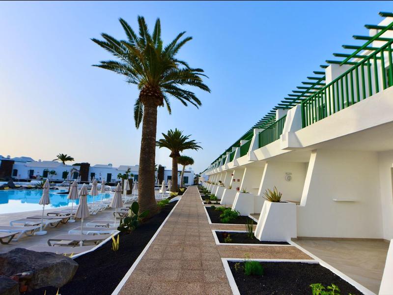 Blick auf einen Poolbereich mit Liegestühlen und einem weißen Hotelgebäude mit grünen Akzenten.