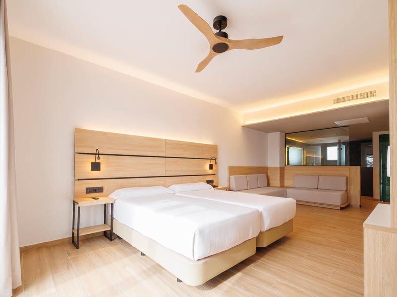 Hell eingerichtetes Hotelzimmer mit zwei Einzelbetten, Holzfußboden und Deckenventilator.