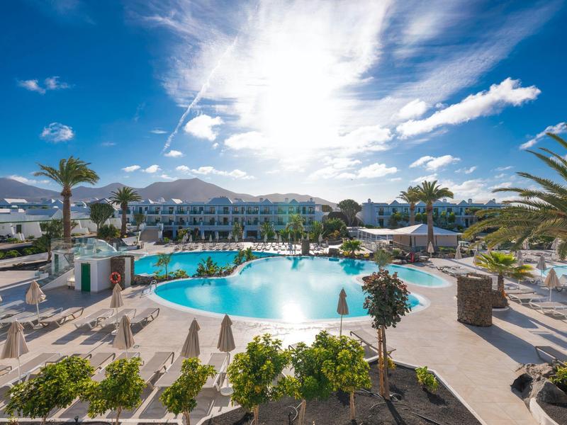 Sonniger Hotelpool mit Palmen und Blick auf Berge im Hintergrund.