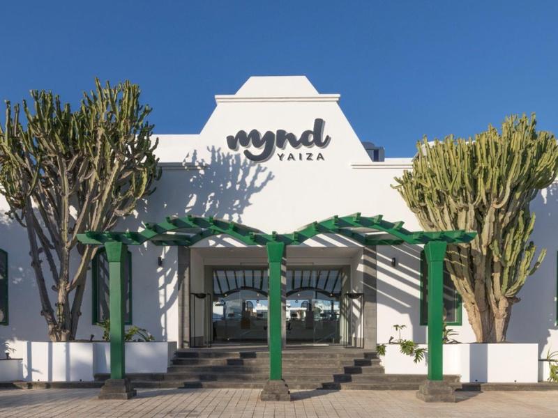 Weißes Gebäude mit grünem Pergola-Eingang und mehreren Kakteen bei klarem Himmel