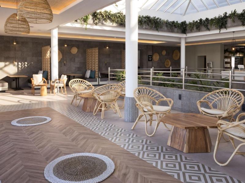 Heller Loungebereich mit Korbsesseln, Holztischen, Pflanzen und geometrischem Bodenmuster.