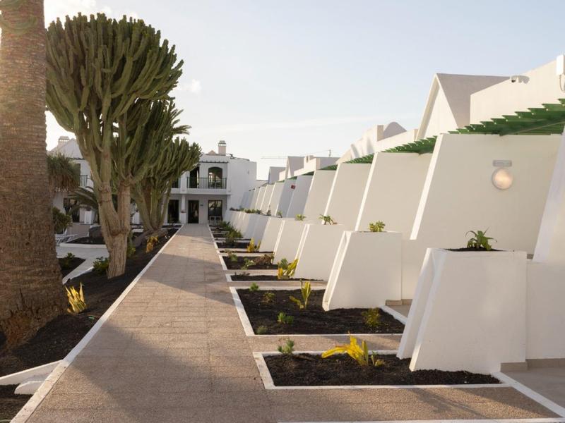 Weißes Terrassenhotel mit Pflanzen und einem breiten Gehweg an einem sonnigen Tag.