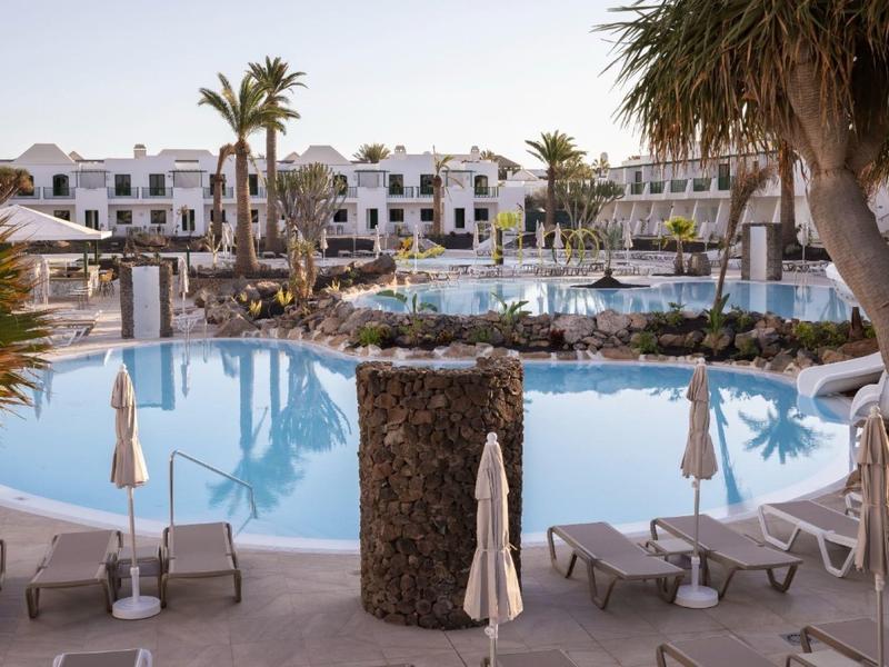 Großer blauer Pool in Resort mit weißen Liegen, Sonnenschirmen, Palmen und weißen Gebäuden im Hintergrund.