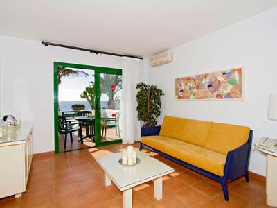 Helles Wohnzimmer mit gelbem Sofa, weißem Tisch und großer Glastür zum Balkon mit Blick aufs Meer.