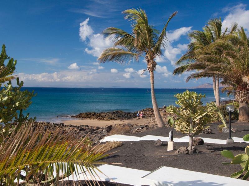 Tropische Küste mit Palmen, schwarzem Vulkansand und klarem blauem Meer unter blauem Himmel.