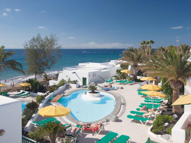 Schwimmbad mit Sonnenschirmen und Liegen, weiße Gebäude, Palmen, Meer und blauer Himmel im Hintergrund.