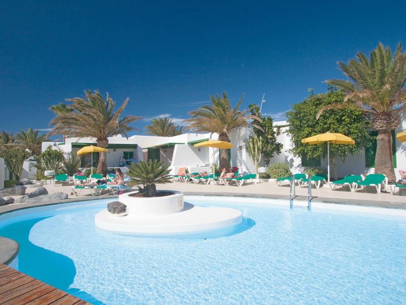 Außenpool mit blauer Wasserfläche, weißen Liegen, gelben Sonnenschirmen und Palmen in hellem Ambiente.