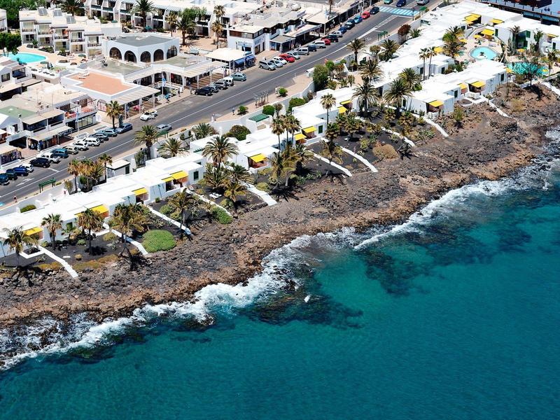 Küstenlinie mit Felsen, blauen Meerwasser und weißen Gebäuden mit Terrassen und Palmen im Hintergrund.