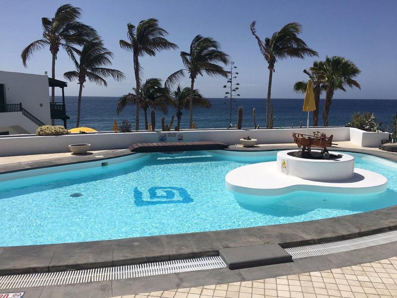 Hellblauer Pool mit runder Insel, Palmen im Hintergrund, Blick aufs Meer bei klarem Himmel.