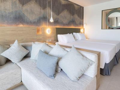 Modernes Schlafzimmer mit zwei Betten, Sofa, vielen Kissen und Holzpaneelwand mit Beleuchtung.