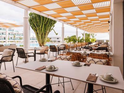 Moderner Außenbereich eines Cafés mit Tischen, Stühlen und orangem Sonnenschutzdach.