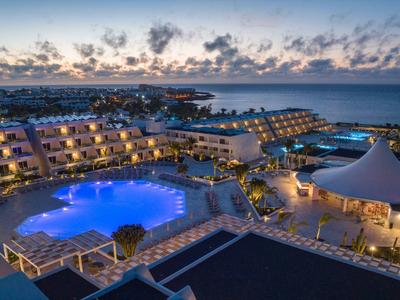 Große Hotelanlage mit beleuchtetem Pool, Terrassen, Meerblick und bewölktem Himmel bei Abenddämmerung.