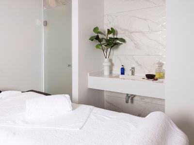 Weißes Schlafzimmer mit Bett, Handtüchern, modernem Waschbecken und Pflanze auf Marmorwand.