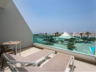 Balkon z dwoma leżakami, stołem i krzesłami, widok na basen i teren hotelu przy czystym niebie.