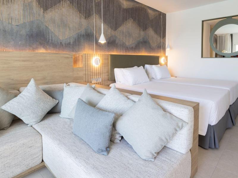 Modernes Schlafzimmer mit zwei Betten, Sofa, vielen Kissen und Holzpaneelwand mit Beleuchtung.