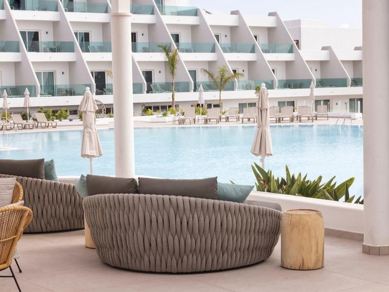 Moderner Sitzbereich mit grauen Sofas, Holzhocker und Blick auf einen Pool vor einem Hotelgebäude.