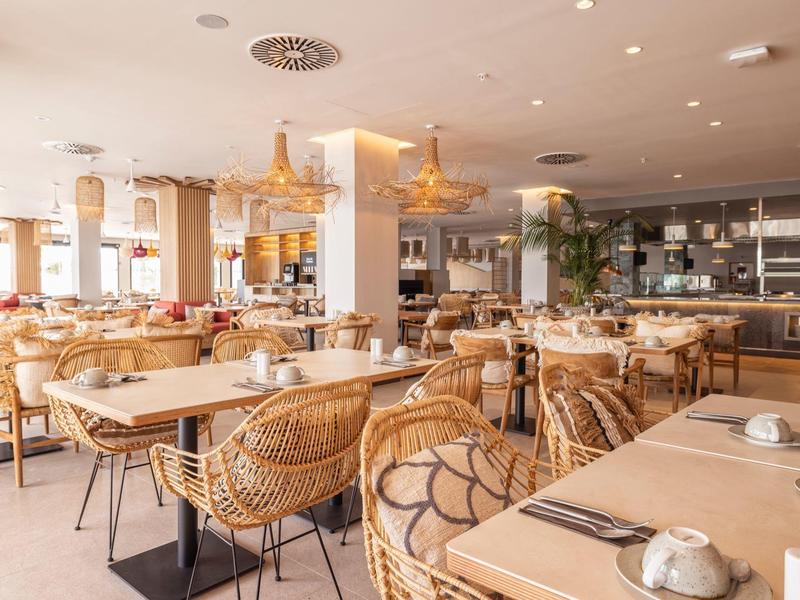 Helles, modernes Restaurant mit Holz- und Rattanmöbeln, großen Fenstern und hellen Lampen an der Decke.