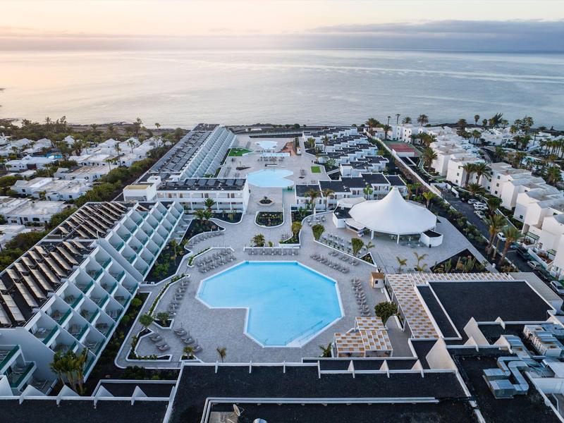 Großer Hotelkomplex mit mehreren Pools nahe der Küste, umgeben von Bäumen und Gebäudezeilen.