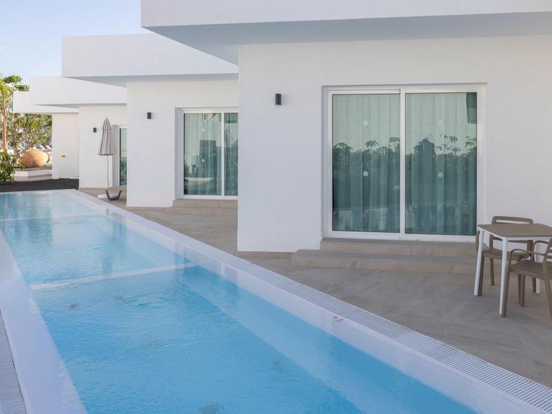 Moderner Außenbereich mit langem, schmalem Pool, weißem Haus und kleiner Sitzgruppe auf grauer Terrasse.