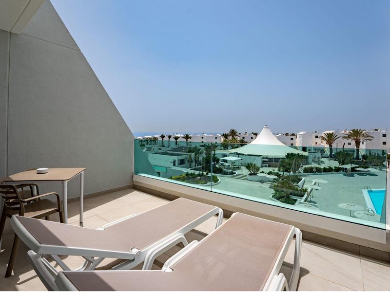 Terrasse mit Liegestühlen und Blick auf Pool und Bungalows unter blauem Himmel.