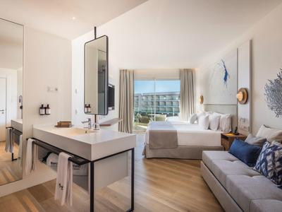 Helles Hotelzimmer mit Doppelbett, Sofa, großem Spiegel und Blick auf grünes Tal durch Fenster.