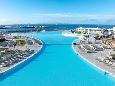 Großer blauer Pool mit Liegestühlen und Blick auf das Meer unter klarem Himmel.
