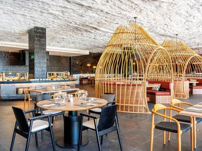 Modernes Restaurant mit Holz- und Rattanmöbeln, runden Tischen und gemütlichen Sitzbereichen.