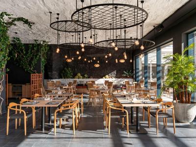 Modernes Restaurant mit Holzmöbeln, hängenden Lampen und großen Fenstern, die Tageslicht hereinlassen.