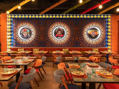 Restaurant mit orangefarbenen Wänden, Holztischen, bunten Stühlen und großen dekorativen Wandbildern.