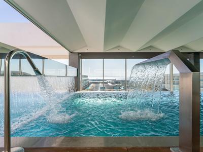 Moderner Hallenpool mit Wasserfall und Edelstahl-Geländer, klares Wasser und große Fenster.