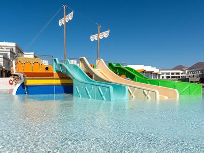 Mehrfarbige Wasserrutschen in einem flachen Pool vor hellen Gebäuden und blauem Himmel.