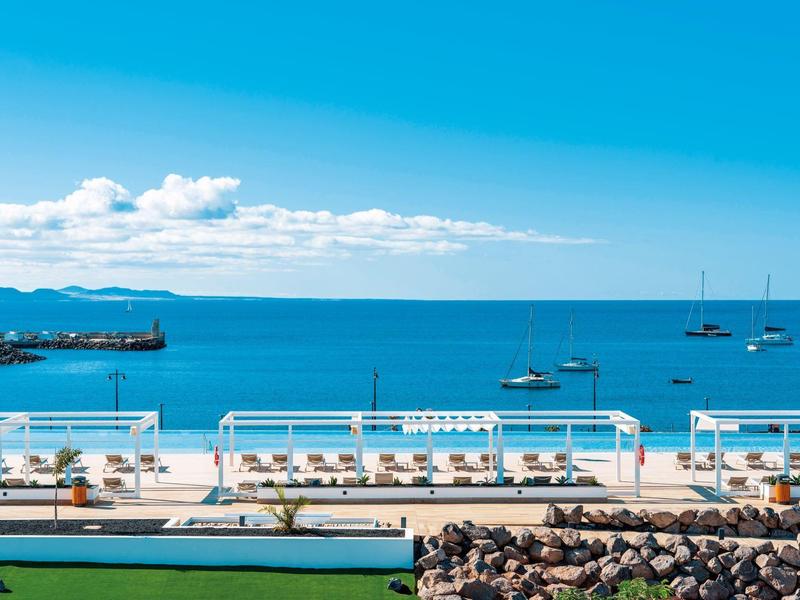Klares Meer und blauer Himmel, Relaxliegen, grüne Wiese, Steine und Yachten im Hafen.