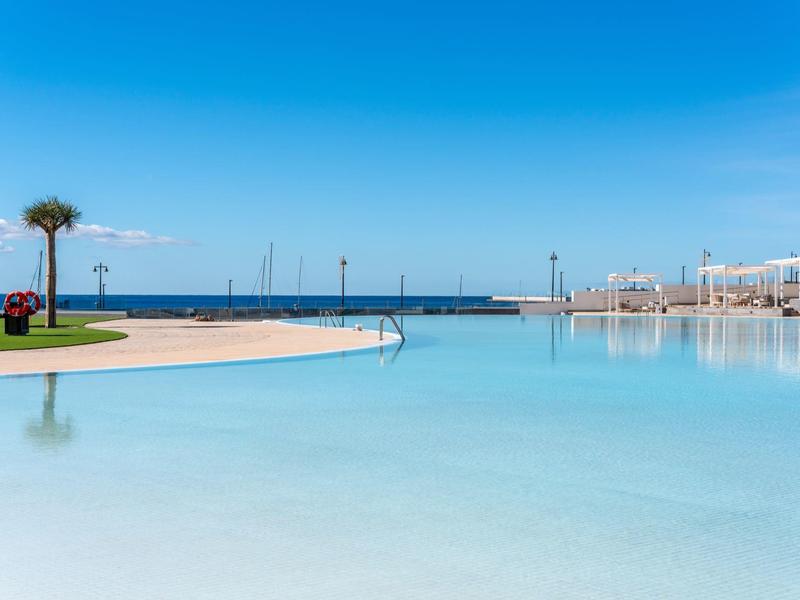 Großer, klarer Pool mit Palmen und Blick aufs ruhige, blaue Meer unter strahlend blauem Himmel.