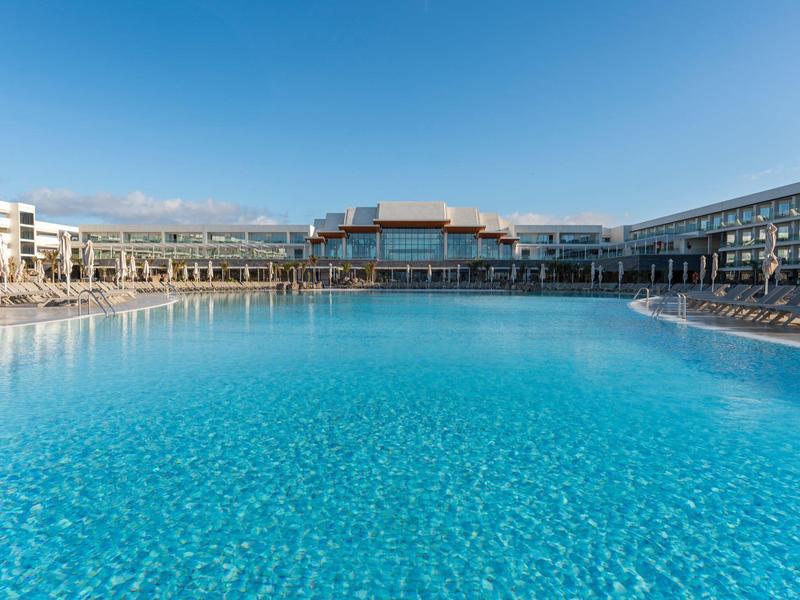 Großer blauer Hotelpool mit Liegestühlen und moderner Architektur bei klarem Himmel.