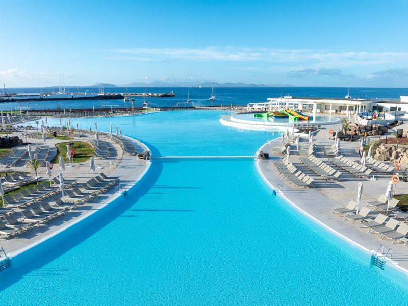 Großer blauer Pool mit Liegestühlen und Blick auf das Meer unter klarem Himmel.