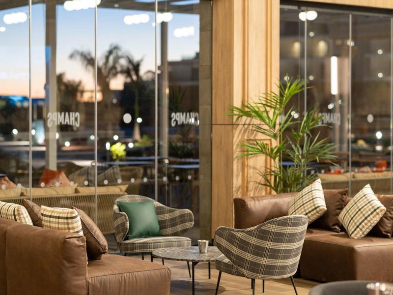 Gemütliche Lounge mit bequemen Sesseln, Sofas, Kissen und Pflanzen vor großen Glasfenstern.