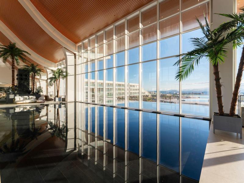 Innenraum mit langem Pool, großen Glasfenstern, Palmen und moderner Architektur bei Tageslicht.