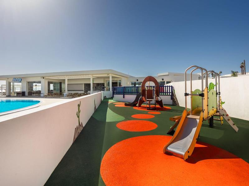 Kleiner Spielplatz mit Rutschen, bunter Gummimatte und angrenzendem Swimmingpool bei klarem Himmel.