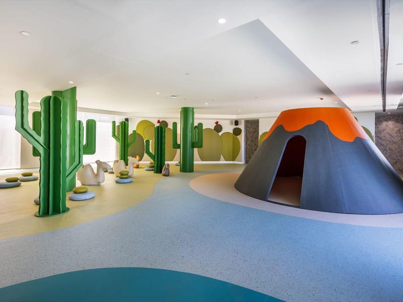 Helle Indoor-Spielhalle mit grünen Kaktussen und einer großen Vulkanrutsche.