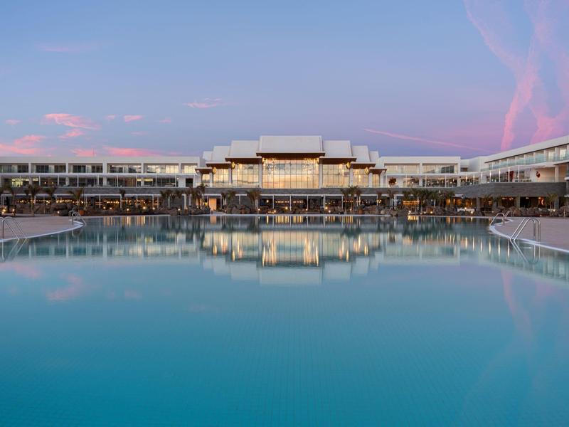Ein großes modernes Hotel mit langgestrecktem Pool und rosa-blauem Abendhimmel.