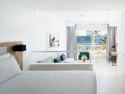 Hell eingerichtetes Hotelzimmer mit großem Bett, Sofa, Balkon und Meerblick bei Tageslicht.